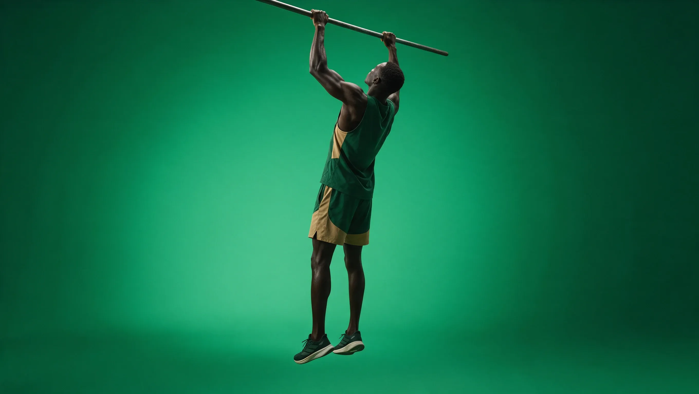 Un athlète suspendu à une barre horizontale par les deux mains, jambes tendues, en tenue verte et sable sur fond vert.